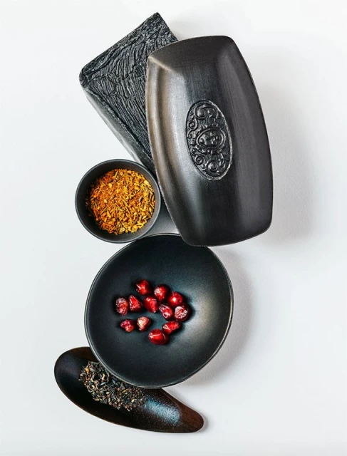Black-themed product photography featuring soap, spices, pomegranate seeds, and tea in bowls against a bright, neutral background.