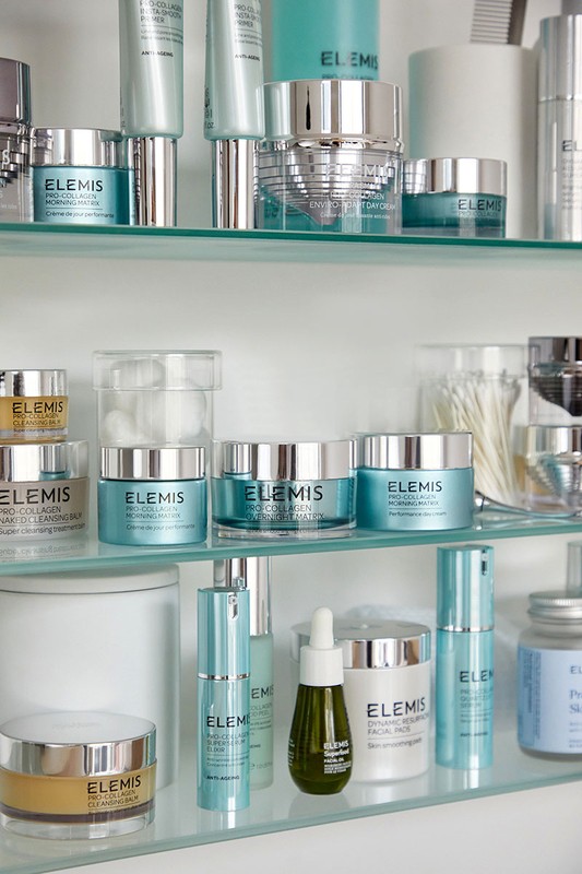 Various Elemis skincare products are displayed on glass shelves, including creams, serums, and cleansers in light blue, white, and silver packaging.