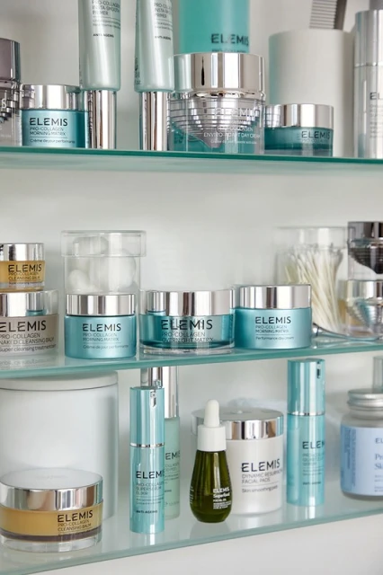 Various Elemis skincare products are displayed on glass shelves, including creams, serums, and cleansers in light blue, white, and silver packaging.