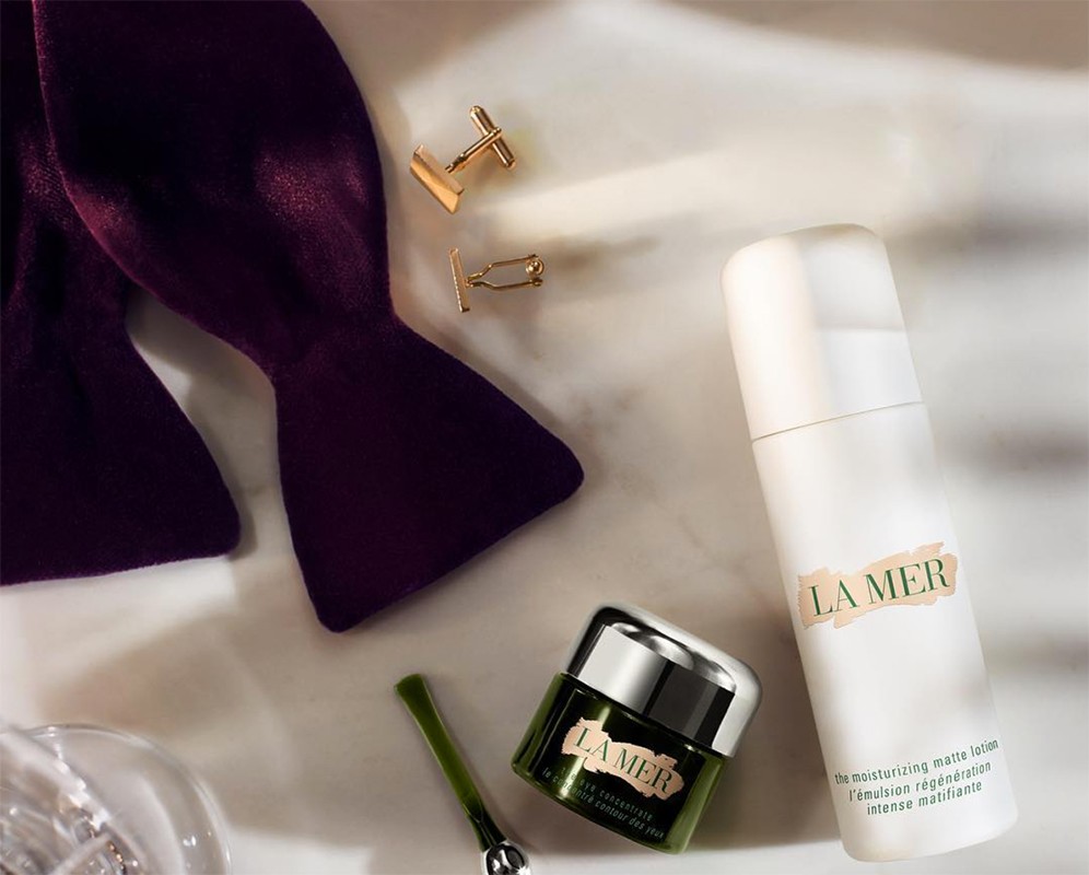 La Mer skincare product arrangement on a white surface featuring a moisturizer, lotion, bow tie, and gold cuff links.