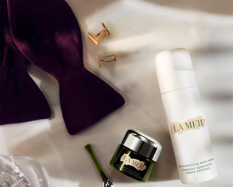 La Mer skincare product arrangement on a white surface featuring a moisturizer, lotion, bow tie, and gold cuff links.