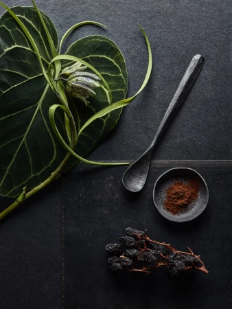 Top-down view of green leaf, fritillary flower, dark spoon, small bowl of spice powder, and dried fruit on a dark stone surface.
