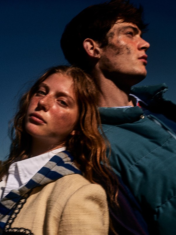 A young woman and man pose outdoors, the woman with wavy ginger hair, wearing a blazer and the man wearing a teal puffer jacket.