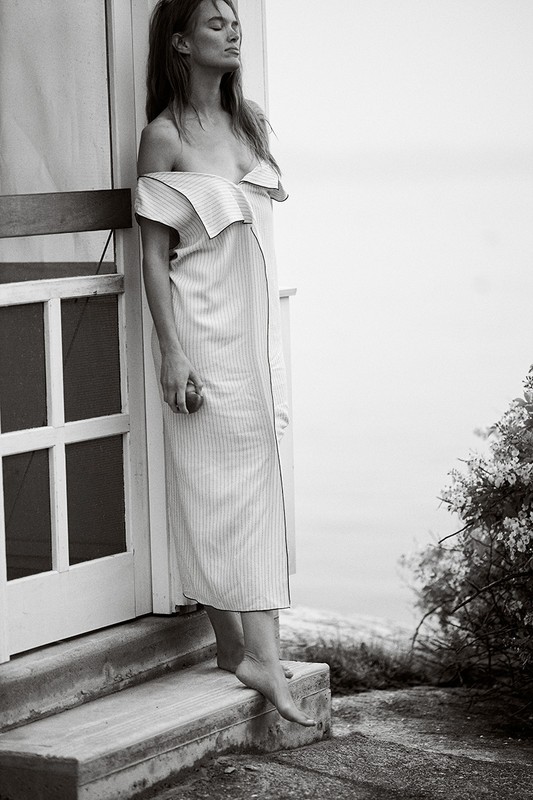 Adult woman in a striped dress stands barefoot by a doorway on concrete steps with a view of the water in a black and white photograph.
