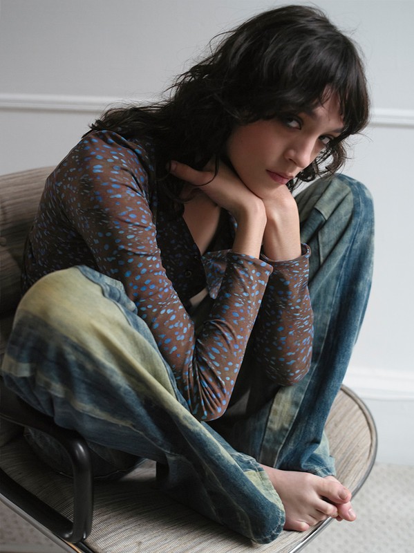 Brunette woman in patterned top and tie-dye jeans sits on a chair indoors with her hands under her chin, looking at the camera.