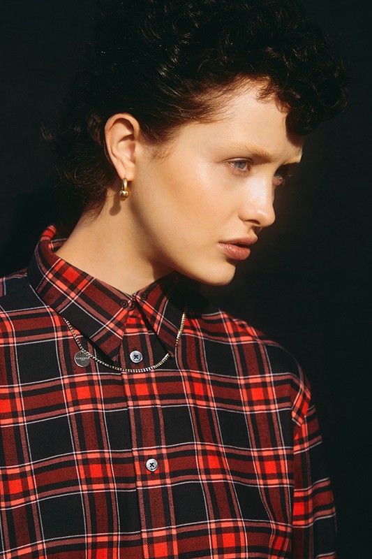 Woman in red and black plaid shirt with gold hoop earrings against black backdrop, captured in a medium shot under bright lighting.