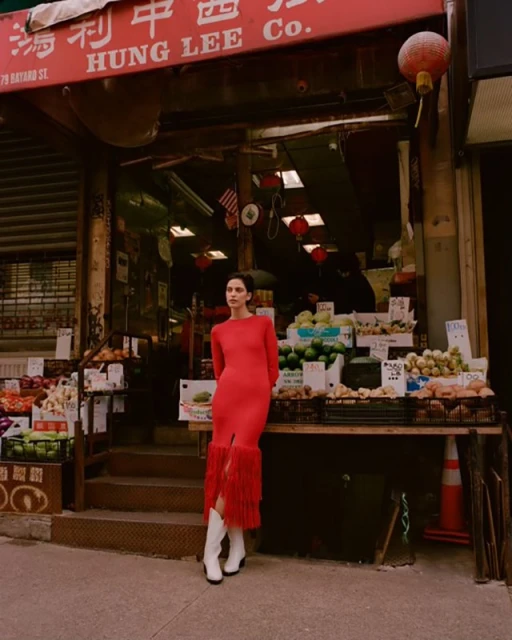 A woman in a red dress and white boots poses outside a store in Chinatown, surrounded by fruit and vegetables on display.