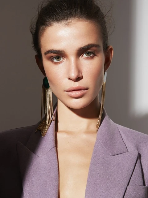 Close-up portrait of a woman wearing a lilac blazer and long, gold tassel earrings against a grey studio backdrop with soft lighting.