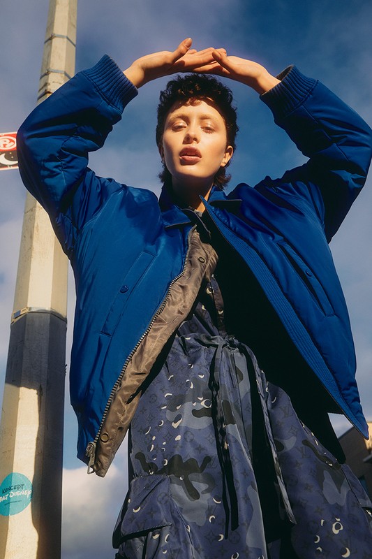 Woman in blue jacket and camouflage dress stands outdoors with hands above head, posing in urban setting with a utility pole.
