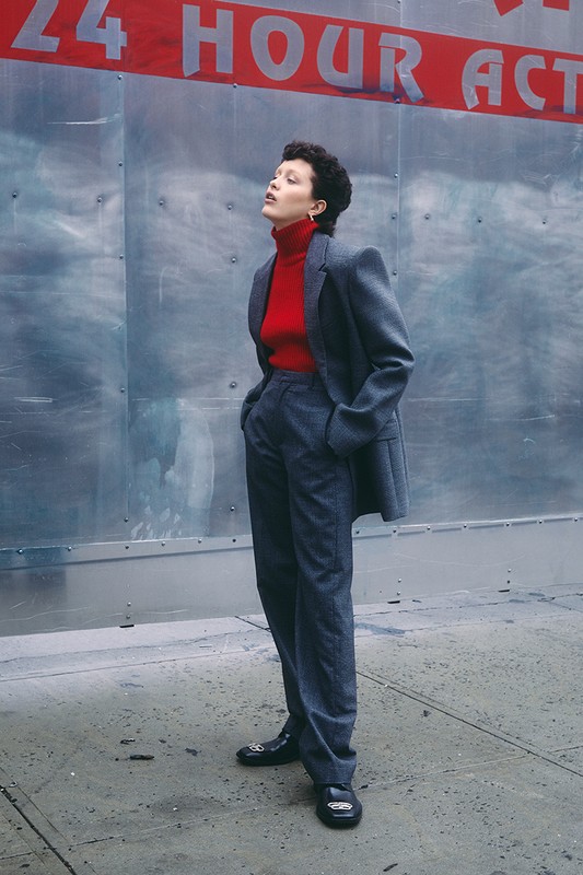 A woman in a grey suit and red turtleneck poses outdoors against an industrial metal wall with a red sign on top of the wall.