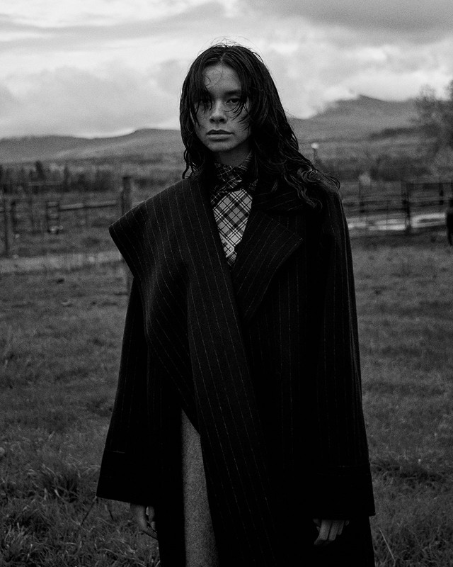 Monochrome portrait of a woman in a coat, standing in a grassy field with a blurred mountain background under an overcast sky.