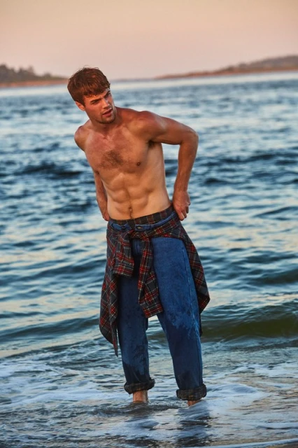 Athletic man standing in ocean tide, shirt tied around waist, ocean backdrop, blue water, tan skin, toned body, casual and relaxed.
