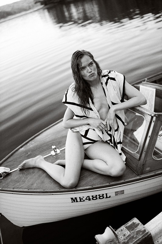 Monochrome portrait of a woman kneeling in a boat wearing a cover-up. Calm water and shoreline in the background. 