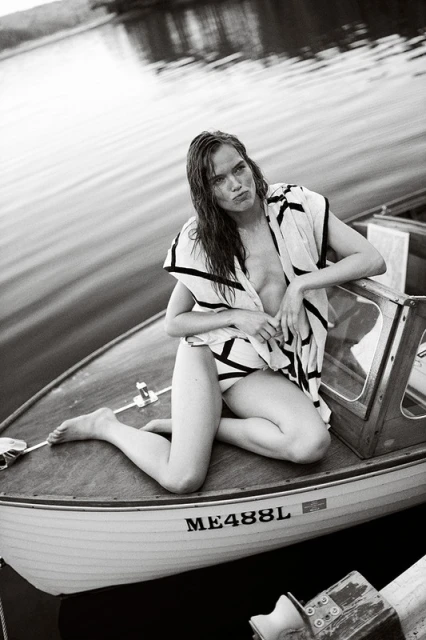 Monochrome portrait of a woman kneeling in a boat wearing a cover-up. Calm water and shoreline in the background. 
