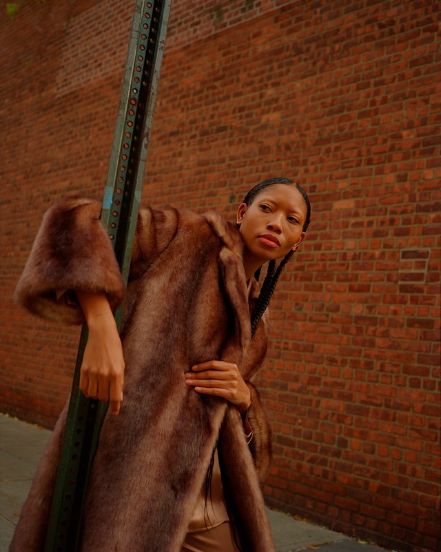 Woman in a fur coat leans against a green pole in front of a brick wall. She has braided hair and is looking at the camera.