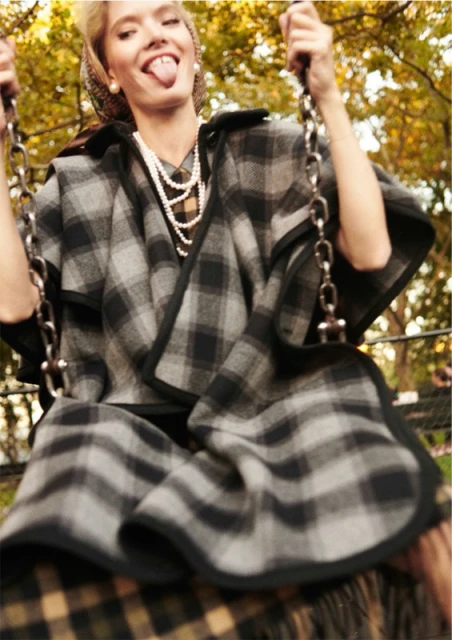 A woman in a plaid coat and headscarf sticks out her tongue while on a swing in an outdoor autumn setting. The photo captures a playful moment.