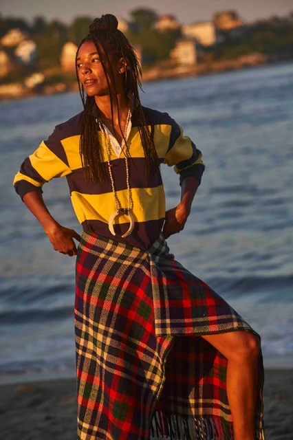 Young woman poses on a beach at sunset wearing a rugby shirt, kilt, and pendant necklace with the ocean and houses in the background.