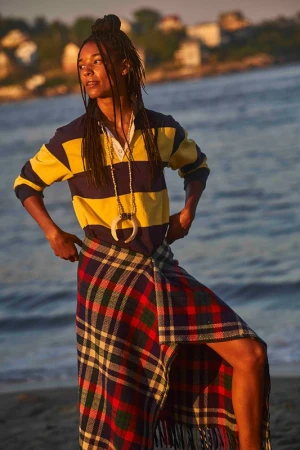 Young woman poses on a beach at sunset wearing a rugby shirt, kilt, and pendant necklace with the ocean and houses in the background.