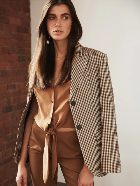 Brunette woman in a brown shirt, blazer, and pants poses indoors against a brick wall. Fashion editorial style portrait.