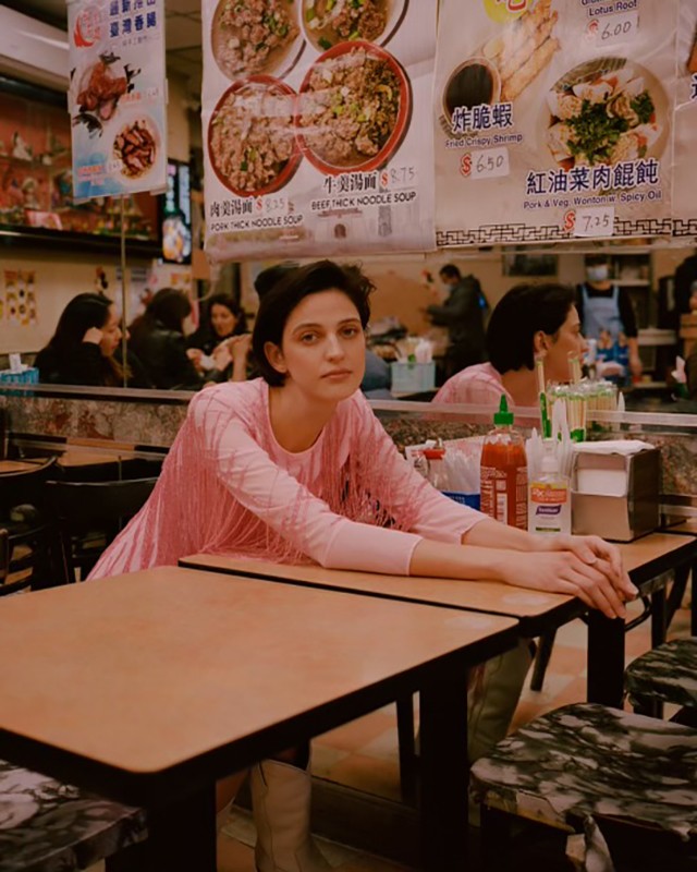 Woman with short dark hair in a pink fringe dress sits at a wooden table in a diner with food menus in the background.