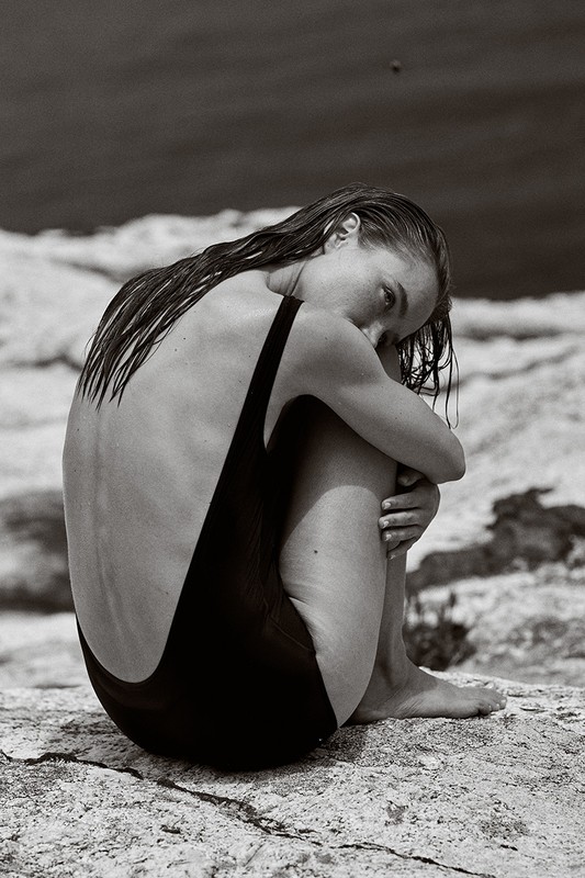 Woman in black swimsuit seated hugging legs on a rock formation in a grayscale image with a blurred background of water.