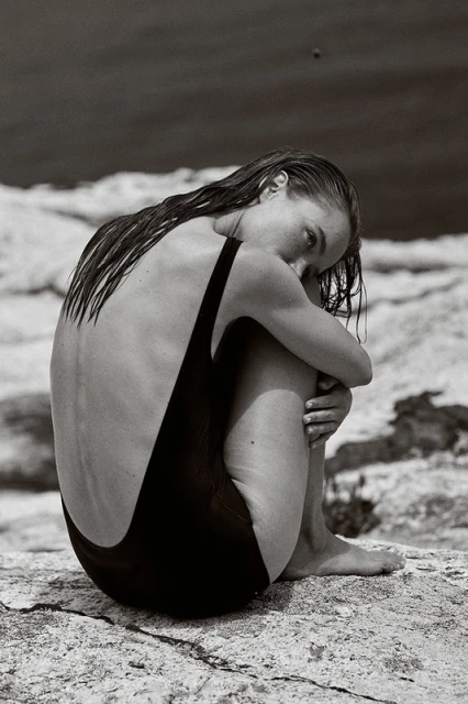Woman in black swimsuit seated hugging legs on a rock formation in a grayscale image with a blurred background of water.
