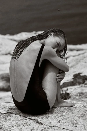 Woman in black swimsuit seated hugging legs on a rock formation in a grayscale image with a blurred background of water.