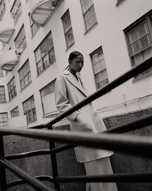 Fashion shot of a woman in a coat leaning against a concrete railing with a building facade visible in the background in black and white.