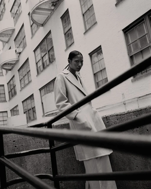 Fashion shot of a woman in a coat leaning against a concrete railing with a building facade visible in the background in black and white.