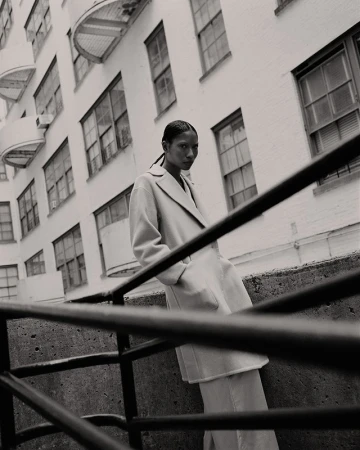 Fashion shot of a woman in a coat leaning against a concrete railing with a building facade visible in the background in black and white.