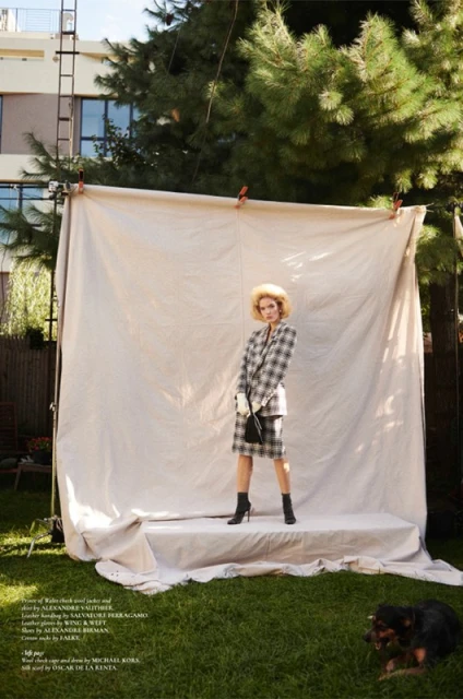 Woman in plaid suit poses with a dog in an outdoor studio setup with a backdrop, ladder, and natural green foliage.