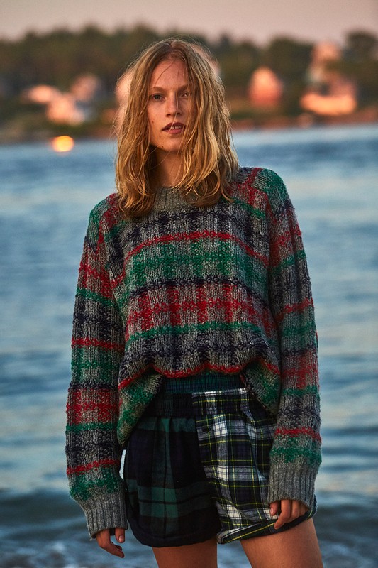 Fashionable woman in knit sweater and plaid shorts posing on a beach against a scenic ocean and sky backdrop at dusk.
