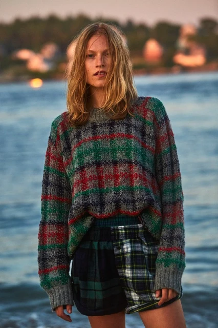 Fashionable woman in knit sweater and plaid shorts posing on a beach against a scenic ocean and sky backdrop at dusk.