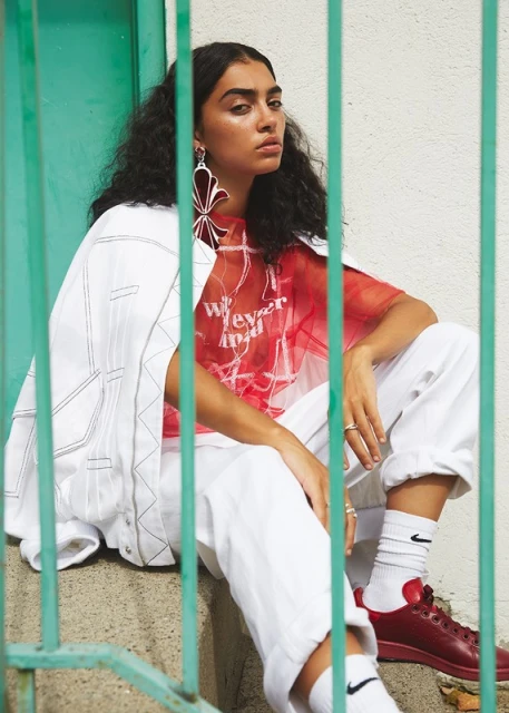 A young adult woman with dark curly hair poses in casual fashion, wearing a white jacket, red top, white pants and red sneakers.