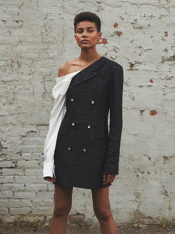 Fashionable woman in one-shoulder dress, a mix of blazer and white shirt, poses confidently against a textured grey brick wall.