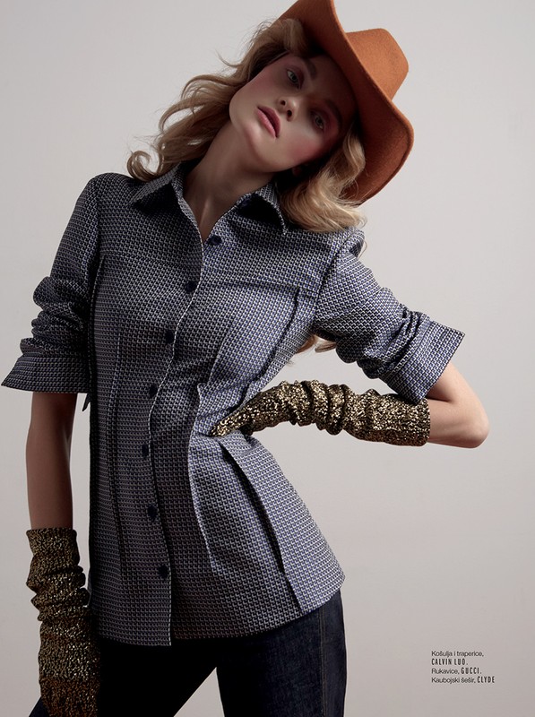 Fashion portrait of a blonde woman in a patterned blue shirt, cowboy hat, and metallic gold gloves, posing against a neutral background.