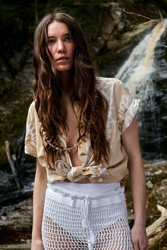 Adult woman poses outdoors by a waterfall wearing a tied patterned short-sleeve shirt and white knit shorts with a beaded necklace.