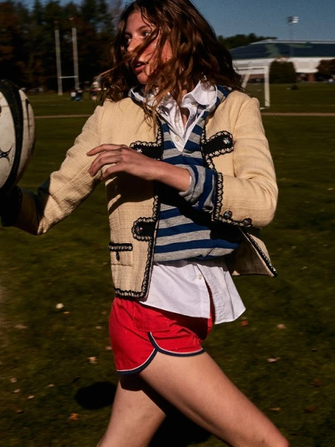 Young woman in athletic wear runs on a grassy field, holding a rugby ball, wearing a cream-colored jacket and red shorts in bright daylight.