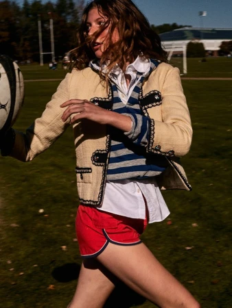 Young woman in athletic wear runs on a grassy field, holding a rugby ball, wearing a cream-colored jacket and red shorts in bright daylight.