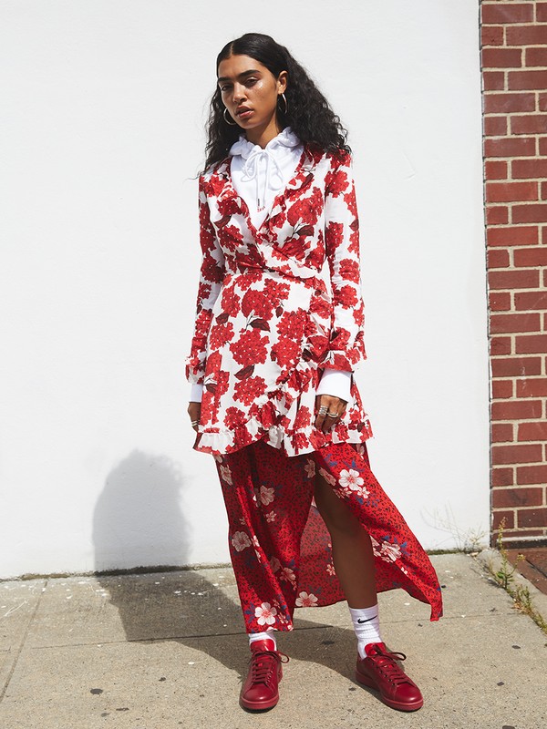 A woman stands in a white and red floral dress, accented by a layered white top and red shoes, against a neutral wall.