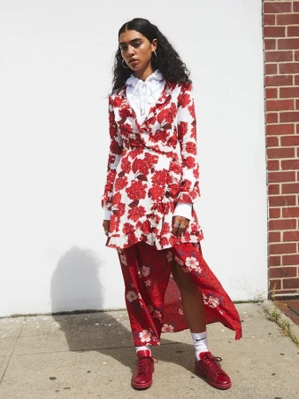 A woman stands in a white and red floral dress, accented by a layered white top and red shoes, against a neutral wall.