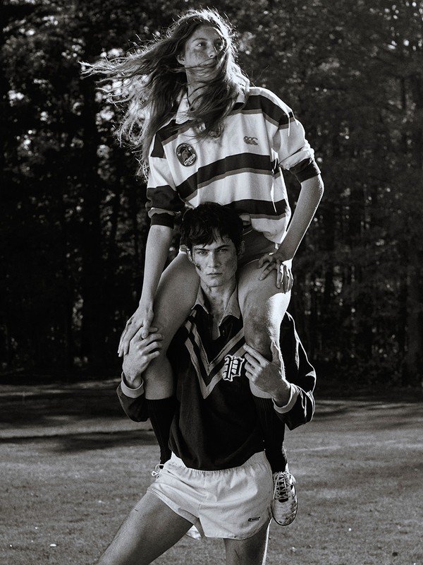 A man carries a woman on his shoulders in a black-and-white shot. Both wear sports jerseys and shorts in an outdoor, grassy setting.