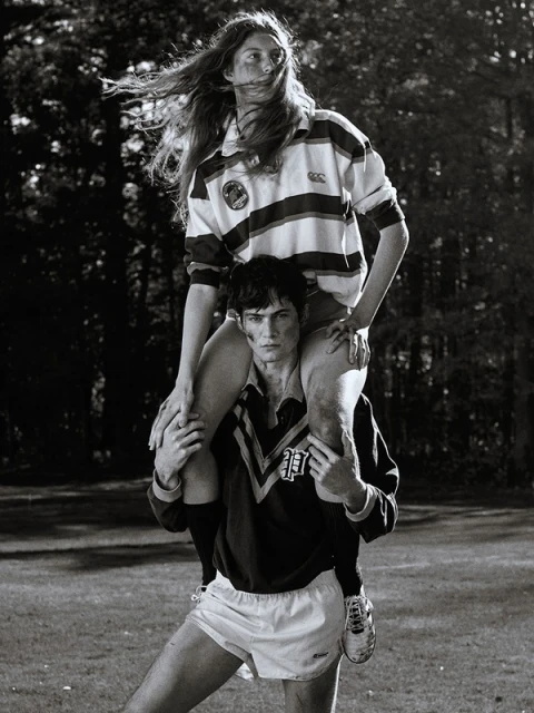A man carries a woman on his shoulders in a black-and-white shot. Both wear sports jerseys and shorts in an outdoor, grassy setting.