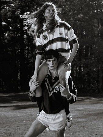 A man carries a woman on his shoulders in a black-and-white shot. Both wear sports jerseys and shorts in an outdoor, grassy setting.