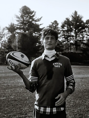 Black and white portrait of a young adult male holding a rugby ball outdoors with trees in the background on a sunny day.