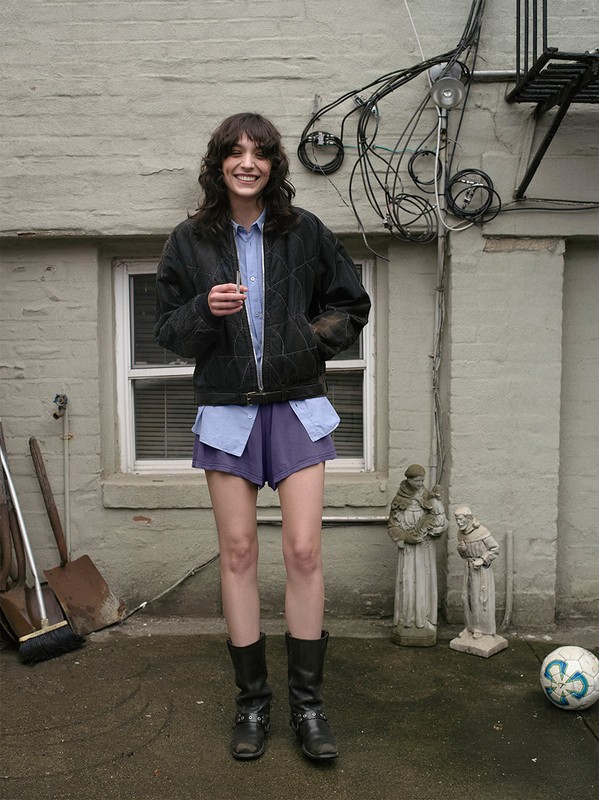 Smiling woman in jacket, shorts, and boots stands in an outdoor setting with garden tools, statues, and a soccer ball nearby.
