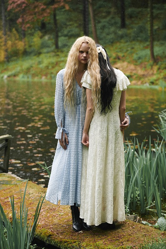 Two women stand on a mossy stone bridge over a pond surrounded by lush greenery in a fashion editorial style outdoor photoshoot.