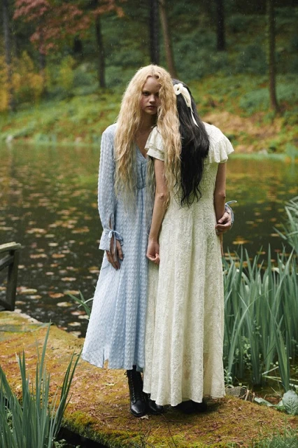 Two women stand on a mossy stone bridge over a pond surrounded by lush greenery in a fashion editorial style outdoor photoshoot.