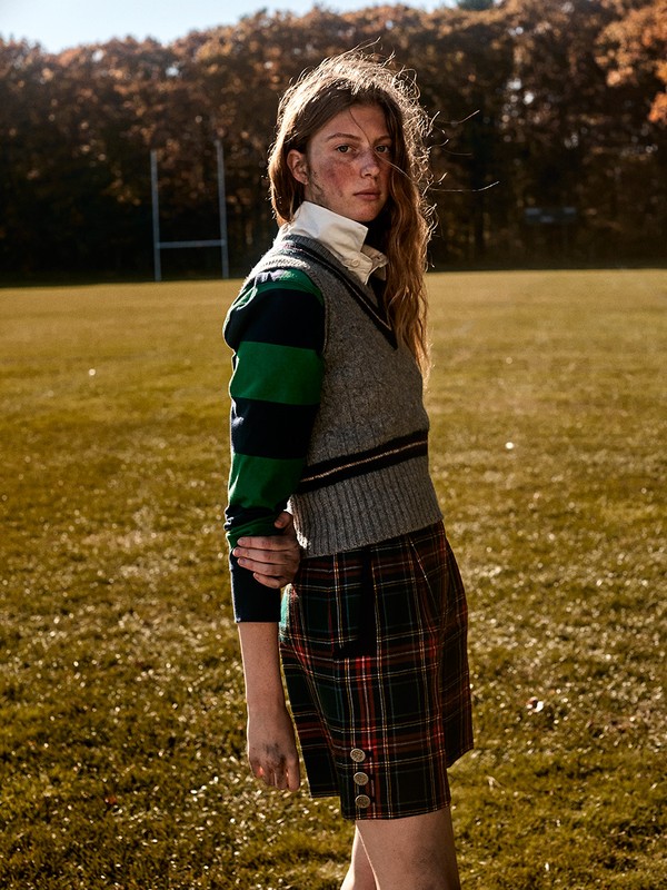 Female posing in preppy style on a field. She wears a sweater vest and plaid shorts, with a field and trees in the background.