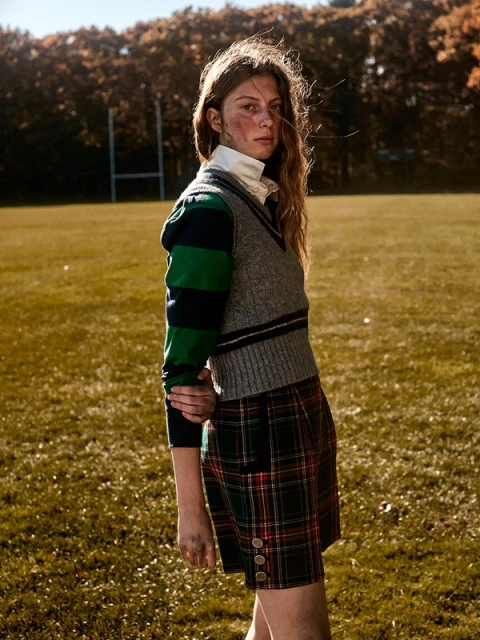 Female posing in preppy style on a field. She wears a sweater vest and plaid shorts, with a field and trees in the background.